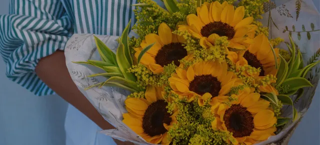 Grande buquê de girassóis com flores amarelas miúdas, nas mãos de uma florista de camisa listrada